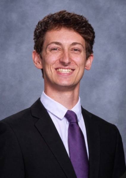 Image 1 of 2 Person smiling wearing a suit in front of a gray wall.